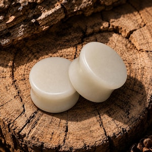 May include: A pair of circular ear plugs with a smooth, polished surface and a pale, off-white color. The plugs feature a concave center section and flared edges, crafted from a solid, opaque material with a consistent, uniform appearance.