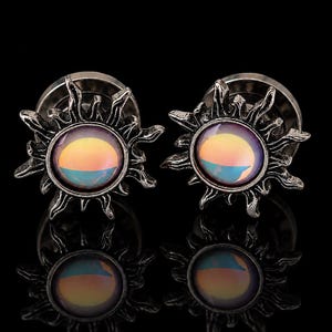 May include: Two silver-coloured ear gauges with a sunburst design. Each gauge has a central, iridescent, circular stone with yellow, orange, and blue hues. The sunburst frame has detailed, pointed rays. The gauges are displayed against a black background.