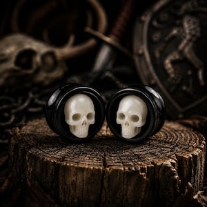 May include: A pair of circular ear plugs featuring a glossy black outer rim and a centered, three-dimensional off-white skull carving. The skull details include defined eye sockets, a nasal cavity, and a realistic jawline set within the black frame.