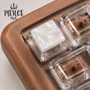 May include: A square white iridescent keycap with a pearlescent finish, mounted on a clear mechanical switch housing with a brown stem. The keycap is set within a dark brown wooden frame. The text PIERCE PLUG with a crown icon is visible.