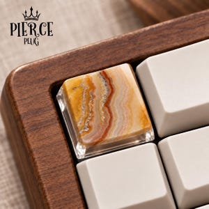 May include: A square mechanical keyboard keycap featuring a polished, natural stone top with swirling bands of amber, orange, and white. The keycap has a clear, transparent base and is positioned within a dark wood keyboard frame next to white keys.