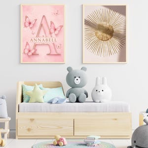 Può includere: Un divano letto in legno chiaro con due cassetti, sormontato da un materasso grigio, un cuscino a forma di stella e cuscini a righe. Due stampe da parete incorniciate mostrano una lettera A rosa con farfalle e un sole dorato. Vari peluche.