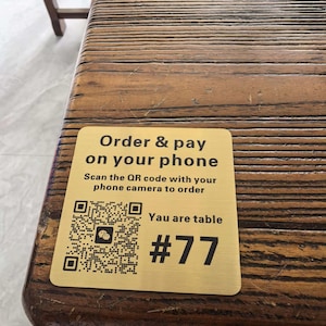 May include: A gold-colored table sign with the text "Order & pay on your phone" and a QR code. The sign also displays "Yau are table #77" on a wooden table.