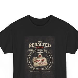 Può includere: T-shirt nera con un design grafico per "The Redacted Club". Il design include il testo "Trust No One - Question Everything", un'immagine di un UFO e "Top Secret". Sono incluse anche le coordinate e la frase "Always Watching - Stay Paranoid".
