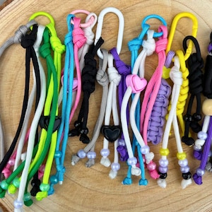 Könnte beinhalten: Eine Sammlung farbenfroher Schlüsselbänder mit verschiedenen Designs. Die Schlüsselbänder weisen eine Vielzahl von Farben auf, darunter Schwarz, Weiß, Rosa, Grün und Lila. Einige Schlüsselbänder haben herzförmige Anhänger, andere Perlen. Sie liegen auf einer Holzoberfläche.