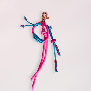 Könnte beinhalten: Ein handgefertigter Makramee-Schlüsselanhänger in Pink und Türkis. Der Schlüsselanhänger hat einen bronzefarbenen Verschluss und dekorative Knoten. Das Design umfasst eine Schlaufe, herabhängende Kordeln und Perlenakzente, was ein einzigartiges Accessoire ergibt.