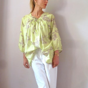May include: A light green blouse with a floral print in a muted grey colour. The blouse has a gathered neckline with a tie detail and 3/4 sleeves.