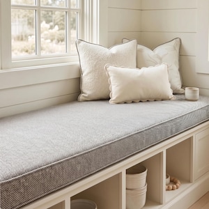 Op de afbeelding: Een lang grijs gestructureerd bankkussen ligt op een witte inbouwkast met open vakken. Drie decoratieve kussens zijn op het kussen gerangschikt: twee vierkante off-white kussens en een kleiner rechthoekig kussen met geschulpte randen.