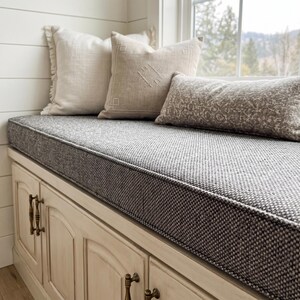 May include: A window seat featuring a grey and white patterned cushion and three decorative pillows. The pillows are in neutral tones, with fringed and embroidered details. The seat is above a white cabinet with brass hardware.