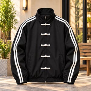 May include: A black track jacket featuring a high collar, a full-length front zipper, and five white toggle-style frog closures. The sleeves are adorned with two parallel white stripes running from the collar to the ribbed cuffs.
