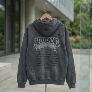 Peut inclure: Un sweat à capuche gris anthracite avec une finition délavée et usée. Le dos présente le texte ARIRANG WORLD TOUR dans une police gothique, suivi d'une liste de villes incluant Londres, Berlin, Paris et d'autres, avec de petits trous.