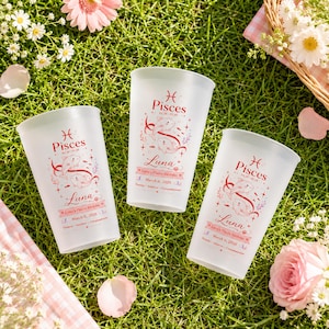 May include: Three frosted plastic cups with a Pisces theme, featuring a red and pink design with the name "Luna" and the date March 6, 2026. The cups are set on green grass with flowers.