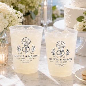 May include: Two clear plastic cups filled with ice, each featuring a dark blue graphic of a seashell above two interlocking rings. The text reads OLIVIA & MASON, JUNE 18, 2027, and ENGAGED BY THE SEA, centered below the rings with a small heart icon.