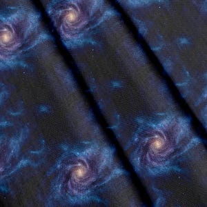 Könnte beinhalten: Ein Stoff mit einem Weltraumdruck, der wirbelnde violette und blaue Spiralgalaxien vor einem tiefschwarzen Hintergrund zeigt. Das Textil weist ein sich wiederholendes Muster aus kosmischen Nebeln und hellen Sternhaufen auf.