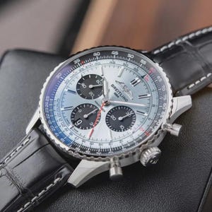 Op de afbeelding: Een Breitling Navitimer chronograaf horloge met een zilveren wijzerplaat, drie zwarte sub-wijzerplaten en een zwarte lederen band. Het horloge heeft een draaibare rekenliniaal-ring, zilverkleurige wijzers en het Breitling 1884 logo op de wijzerplaat.