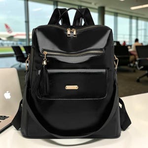 Könnte beinhalten: Ein schwarzer, glänzender Rucksack mit goldfarbenen Reißverschlüssen und Beschlägen. Die Tasche hat eine Vordertasche, eine Quaste und verstellbare Schultergurte. Der Rucksack steht auf einer weißen Oberfläche.