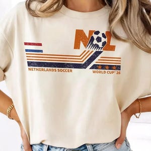 Könnte beinhalten: Cremefarbenes T-Shirt mit einem Grafikdesign zum Thema niederländischer Fußball. Das Design umfasst die Buchstaben "NOL", einen Fußball und den Text "NETHERLANDS SOCCER WORLD CUP '26" in Orange, Blau und Rot.