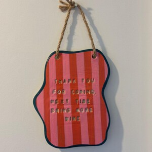 May include: A decorative sign with a unique shape and a rope hanger. The sign features a pink and red striped pattern with the words "THANK YOU FOR COMING, NEXT TIME BRING MORE WINE" in gold lettering. The edges are outlined in dark blue.