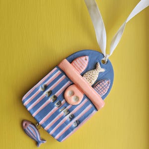 Puede incluir: Una etiqueta de cerámica de Sardinas con un diseño de rayas azules y rosas, con un pequeño dije de pez. La etiqueta tiene la palabra "Sardinas" escrita. Se adjunta una cinta blanca para colgar. El fondo es amarillo.