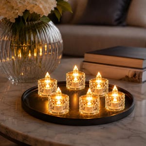 May include: A set of six battery-operated tea light candles with clear, faceted glass holders arranged on a circular black tray. Each candle features a warm, glowing flame-shaped LED bulb that emits a soft light, creating a bright display.