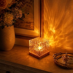 May include: A square glass lamp with a textured, rippled surface that emits a warm, golden light. The lamp sits on a wooden base and casts intricate, wave-like patterns of light onto the adjacent wall, creating a glowing, ambient effect.