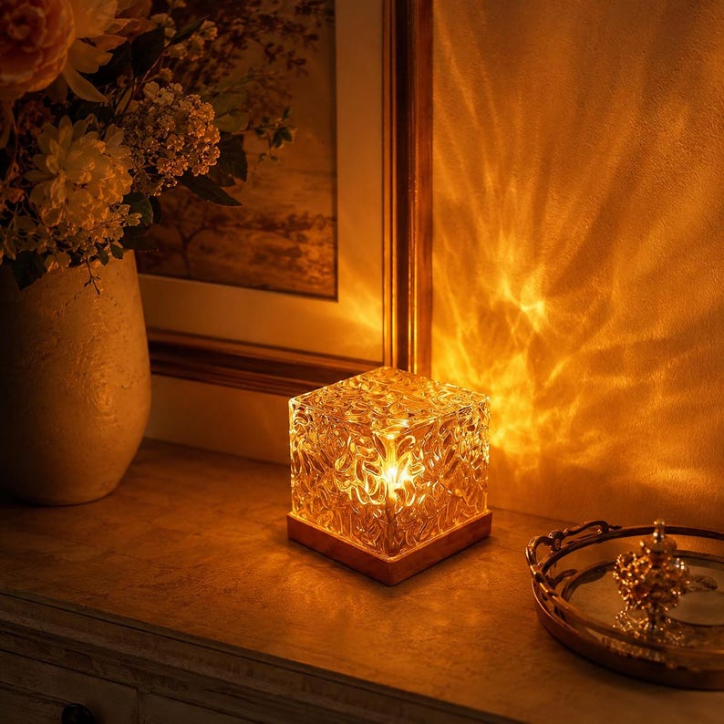 May include: A square glass lamp with a textured, rippled surface that emits a warm, golden light. The lamp sits on a wooden base and casts intricate, wave-like patterns of light onto the adjacent wall, creating a glowing, ambient effect.