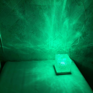 May include: A square-shaped lamp with a textured, translucent body that emits a vibrant green light. The lamp sits on a flat, light-colored base and is connected to a thin white power cord, casting a bright green glow onto the surrounding area.
