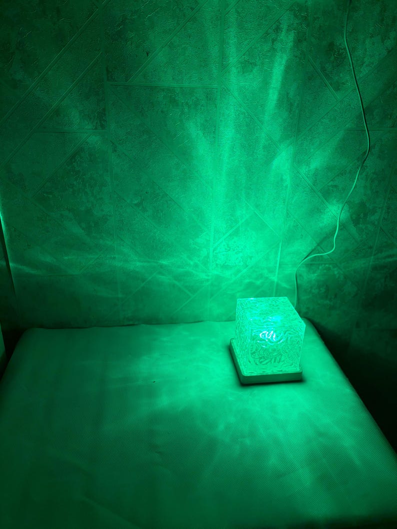 May include: A square-shaped lamp with a textured, translucent body that emits a vibrant green light. The lamp sits on a flat, light-colored base and is connected to a thin white power cord, casting a bright green glow onto the surrounding area.