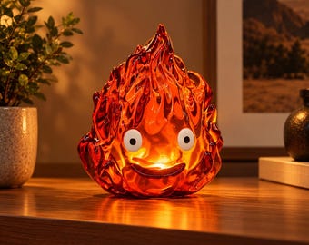 May include: A translucent orange and red flame-shaped lamp with a smooth, glossy finish. The lamp features two round white eyes with black pupils and a wide, curved black mouth, creating a character-like appearance with internal light.