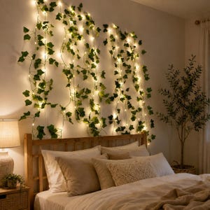 Puede incluir: Una guirnalda de enredaderas de hiedra verde artificial entrelazadas con luces LED de color blanco cálido, colgada verticalmente contra una pared sobre un cabecero de madera. El follaje presenta formas de hojas realistas y luz suave.