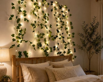 May include: A string of artificial green ivy vines intertwined with warm white LED fairy lights, hanging vertically against a wall above a wooden headboard. The foliage features realistic leaf shapes and the lights emit a soft, ambient glow.