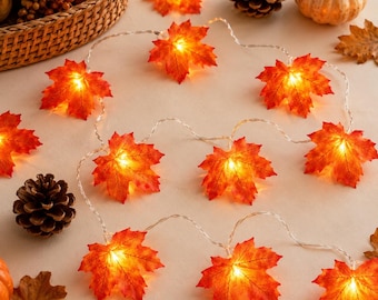 May include: A string of LED fairy lights featuring artificial orange and red maple leaves. Each leaf is illuminated from within, creating a warm glow. The light strand is arranged on a surface alongside pinecones, small pumpkins, and dried leaves.