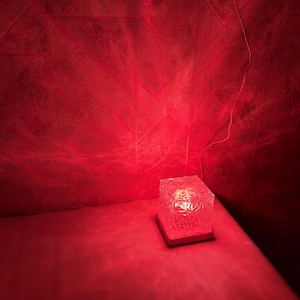 May include: A square, translucent lamp with a textured, crystalline surface that emits a vibrant red light. The lamp sits on a flat, square base and is connected to a thin white power cord extending from the side of the light fixture.
