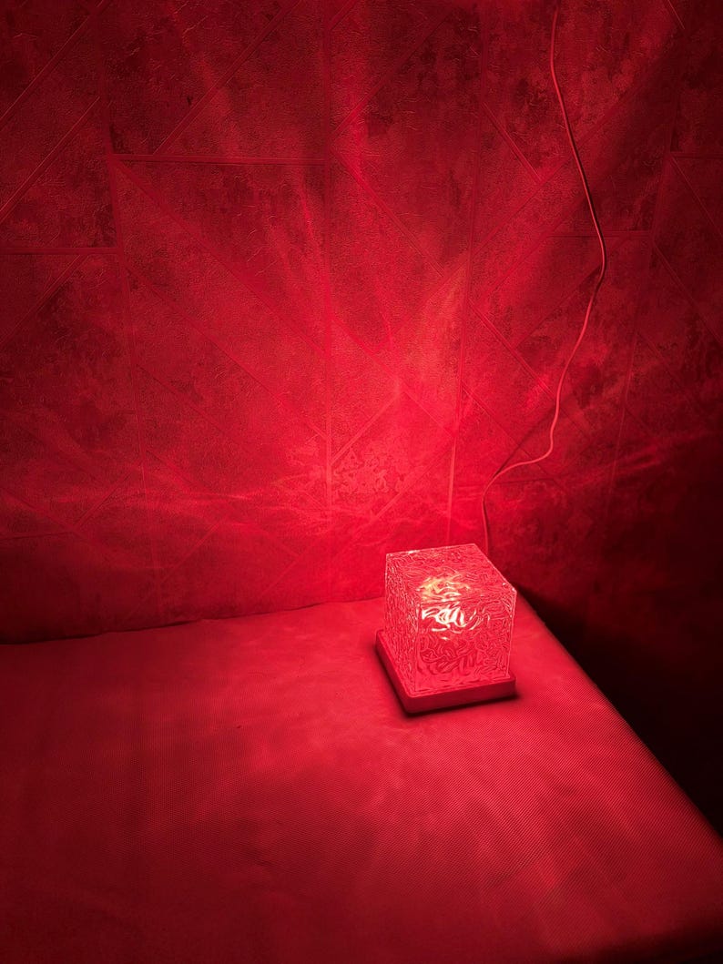 May include: A square, translucent lamp with a textured, crystalline surface that emits a vibrant red light. The lamp sits on a flat, square base and is connected to a thin white power cord extending from the side of the light fixture.