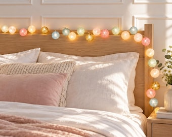 May include: A string of decorative globe lights featuring spherical shades in various pastel colors including pink, blue, yellow, and white. The lights are illuminated, casting a warm glow across the wooden headboard and the bedding below.