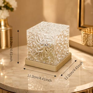 May include: A clear, cube-shaped glass lamp with a textured, rippled surface, set on a square gold-toned base. The lamp measures 11.2 cm or 4.41 inches in height, width, and depth, as indicated by the visible measurement labels.