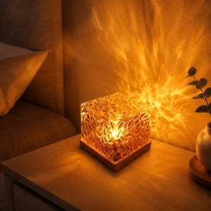 May include: A square glass lamp with a textured, rippled surface that emits a warm, golden light. The lamp sits on a flat wooden base, casting intricate, flame-like light patterns onto the surrounding wall and surface.