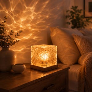 May include: A square-shaped table lamp with a textured, crystalline glass shade that emits a warm, golden light, creating rippling patterns on the wall. The lamp sits on a flat, light-colored wooden base and features a visible internal bulb.