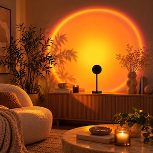 May include: A black sunset projection lamp stands on a wooden sideboard, casting a large, circular, vibrant orange and yellow light onto the wall. The lamp features a thin vertical stem and a small, circular head that emits the glow.