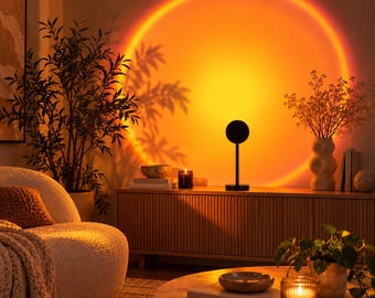 May include: A black sunset projection lamp stands on a wooden sideboard, casting a large, circular, vibrant orange and yellow light onto the wall. The lamp features a thin vertical stem and a small, circular head that emits the glow.