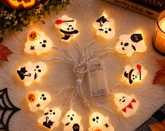 May include: A string of LED lights featuring white ghost-shaped covers with various black and orange Halloween-themed graphics, including pumpkins, cats, bats, and tombstones, connected by a clear wire to a rectangular battery pack.