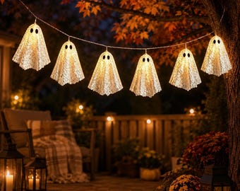 May include: A string of six illuminated ghost-shaped decorations hanging from a tree branch. Each ghost is crafted from white lace fabric with two dark oval eyes, glowing from within to emit a warm light against the dark evening outdoor setting.