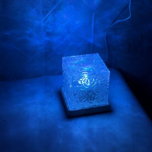 May include: A square-shaped lamp with a textured, translucent glass body that emits a vibrant blue light. The cube rests on a flat, square base and features a rippled, water-like surface pattern that creates dynamic light reflections.