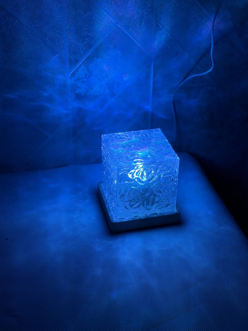 May include: A square-shaped lamp with a textured, translucent glass body that emits a vibrant blue light. The cube rests on a flat, square base and features a rippled, water-like surface pattern that creates dynamic light reflections.