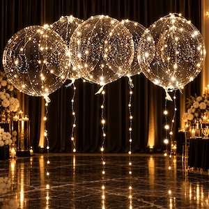 May include: Five large, transparent, spherical balloons, each illuminated by a delicate string of warm white LED lights wrapped around the exterior and trailing down from the base, creating a glowing, starry effect in a dark setting.