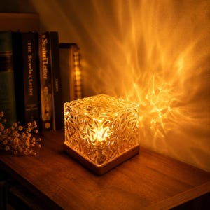 May include: A square glass lamp with a textured, rippled surface that emits a warm, golden light. The lamp sits on a simple wooden base, casting intricate, wave-like patterns of light onto the adjacent wall and surrounding area.