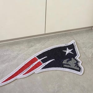 Może przedstawiać: Naszywka z tkaniny z logo New England Patriots, zaprojektowana z ciemnoniebieskim tłem, srebrnym profilem twarzy, białą gwiazdą oraz czerwonymi i białymi paskami, całość wykończona białą obwódką ułatwiającą przymocowanie do odzieży.