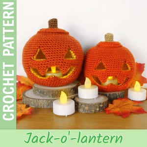 Puede incluir: Dos calabazas Jack-o'-lantern de ganchillo naranja con caras talladas y tallos marrones, cada una iluminada por una pequeña vela. Las calabazas están sobre rodajas de madera, rodeadas de hojas de otoño y velas que funcionan con pilas. El texto "Jack-o'-lantern" está en la parte inferior.