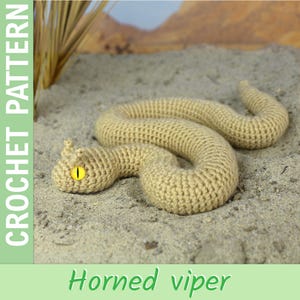 Puede incluir: Una víbora cornuda de ganchillo en hilo beige, con ojos amarillos, descansando sobre arena. La serpiente está en una pose curvada, con el texto "CROCHET PATTERN" y "Horned viper" visible.