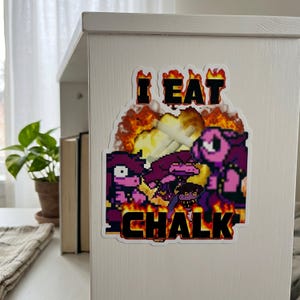 May include: A rectangular sticker featuring pixel art of a purple character with a jagged mouth and glowing eyes, surrounded by bright orange and yellow flames. The text I EAT CHALK is printed in bold black letters against the fiery background.